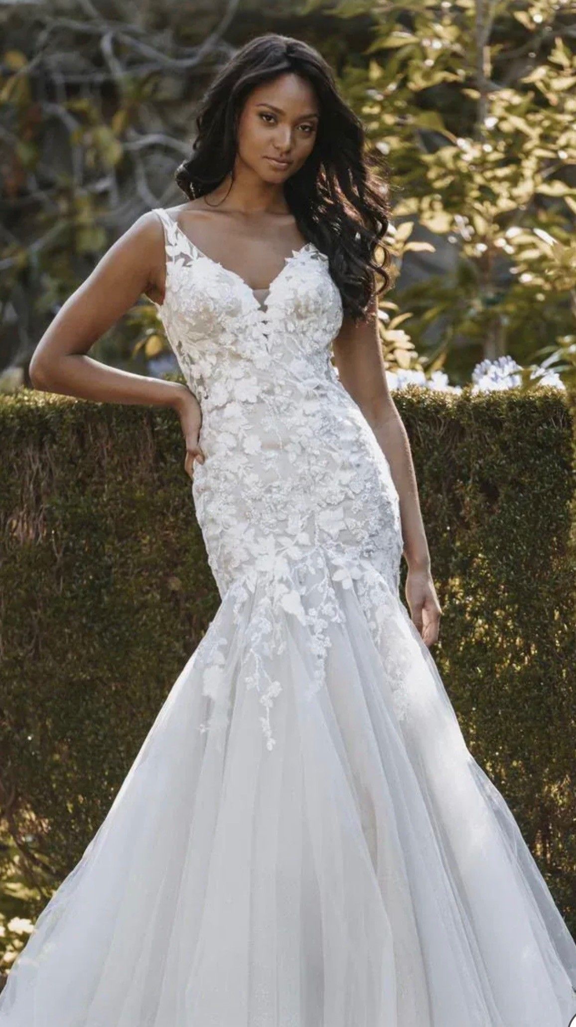 A person wearing a white, V-neck wedding dress with floral lace detailing, standing outdoors in front of a hedge.