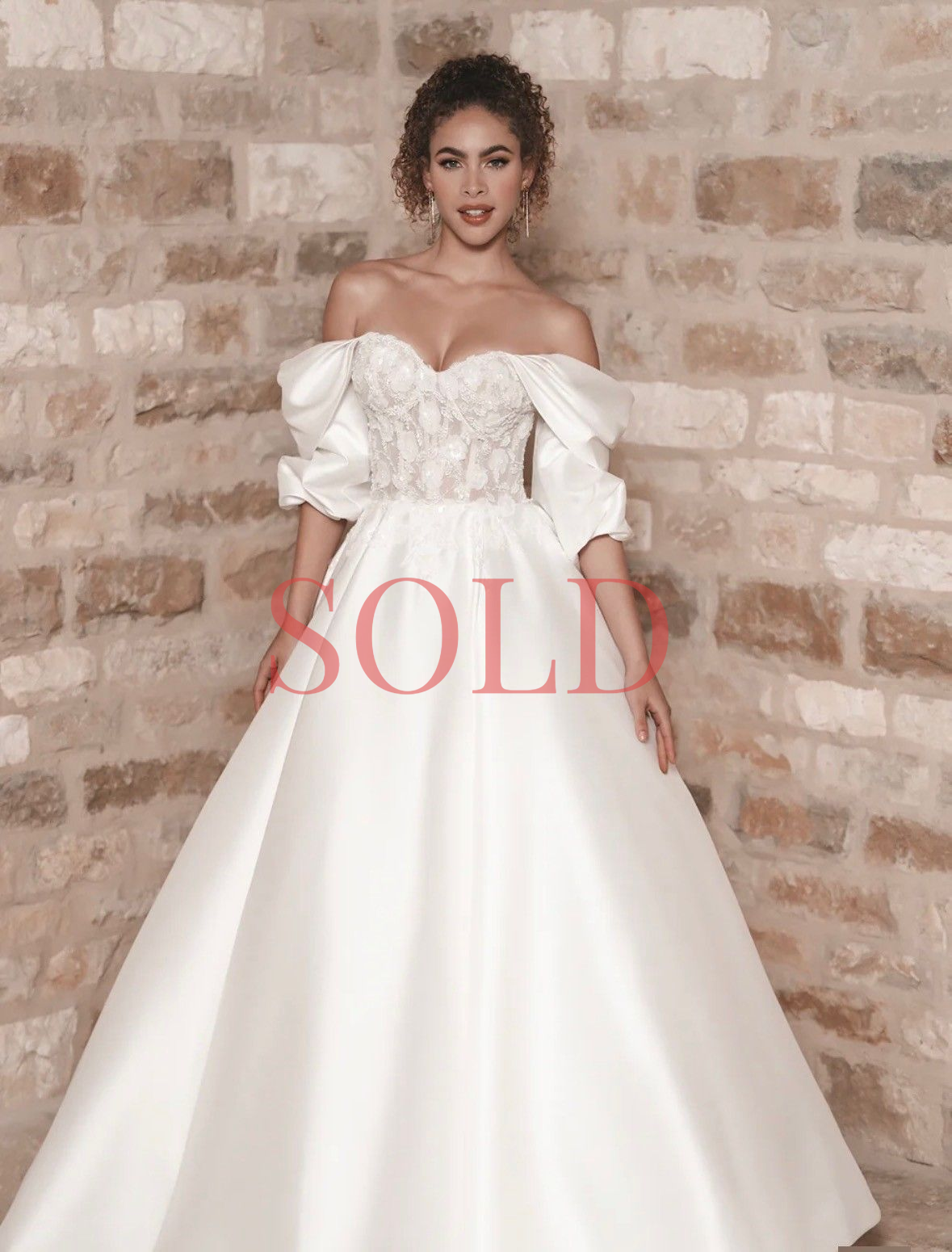 A person wearing an off-the-shoulder, white A-line wedding gown with a floral lace bodice, standing against a brick wall.