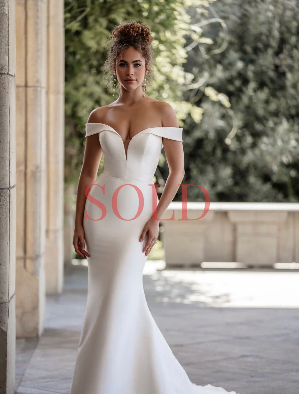 A person wearing a minimalist, off-the-shoulder white mermaid wedding dress with a deep V-neckline, standing outdoors.