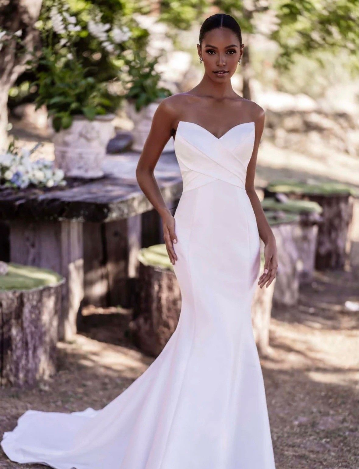 A model wearing a strapless, floor-length white satin mermaid wedding dress, standing in a natural outdoor setting.