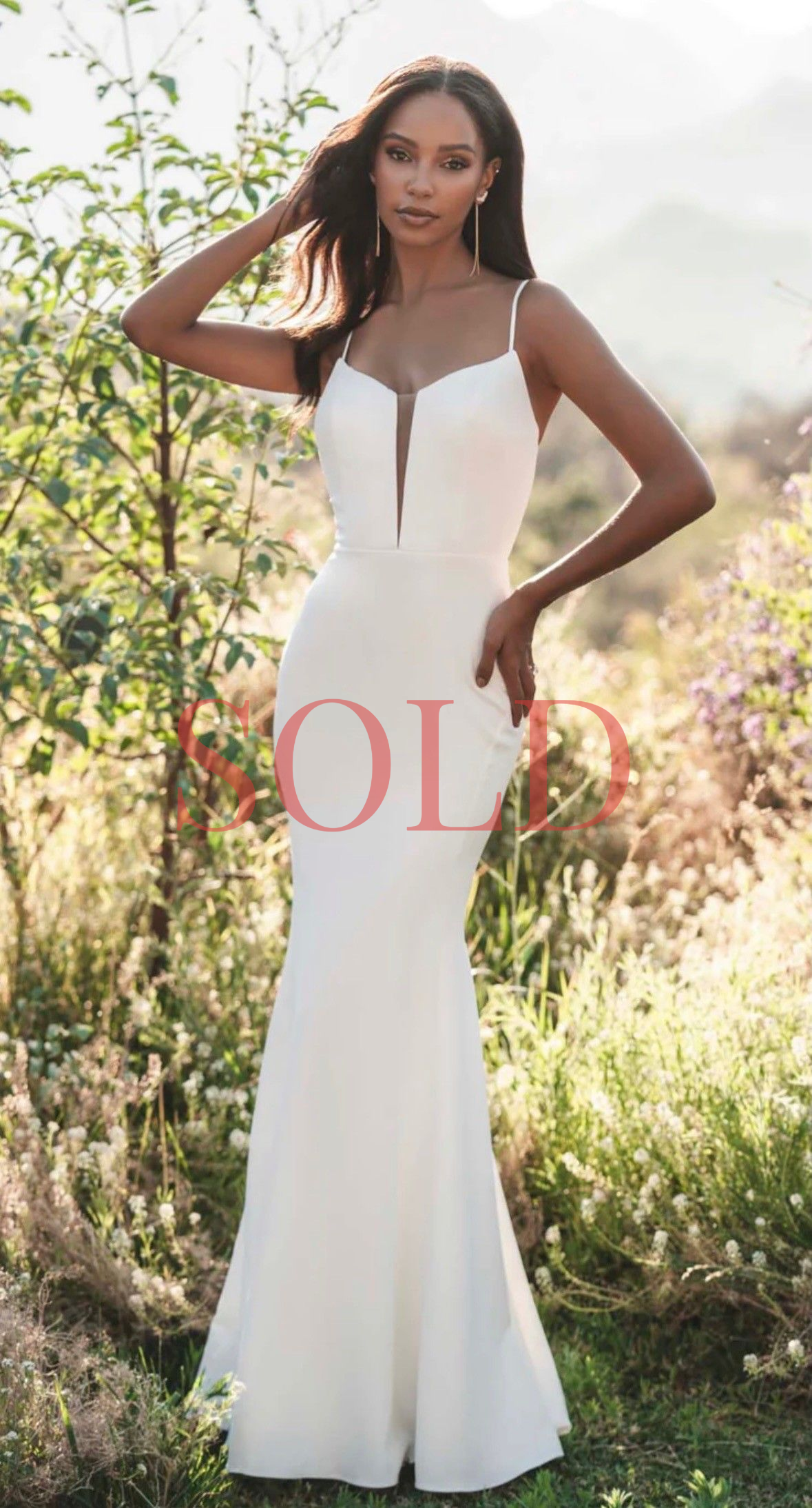 A person wears a form-fitting, sleeveless white floor-length gown with a plunging neckline in an outdoor, grassy setting.
