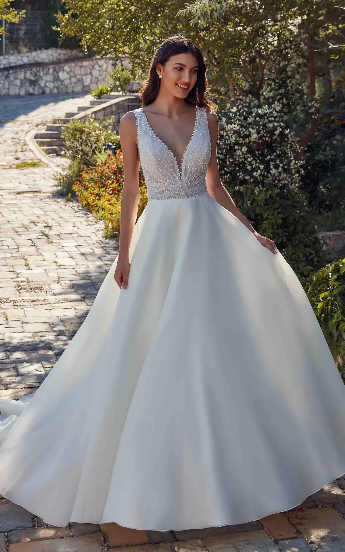 A person wearing a strapless, floral lace mermaid wedding dress with a long train, standing outdoors by a river.