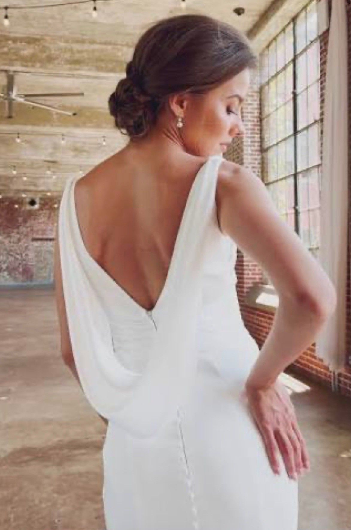 A person wearing a white, backless wedding gown with a draped cowl design, standing in a rustic, light-filled venue.