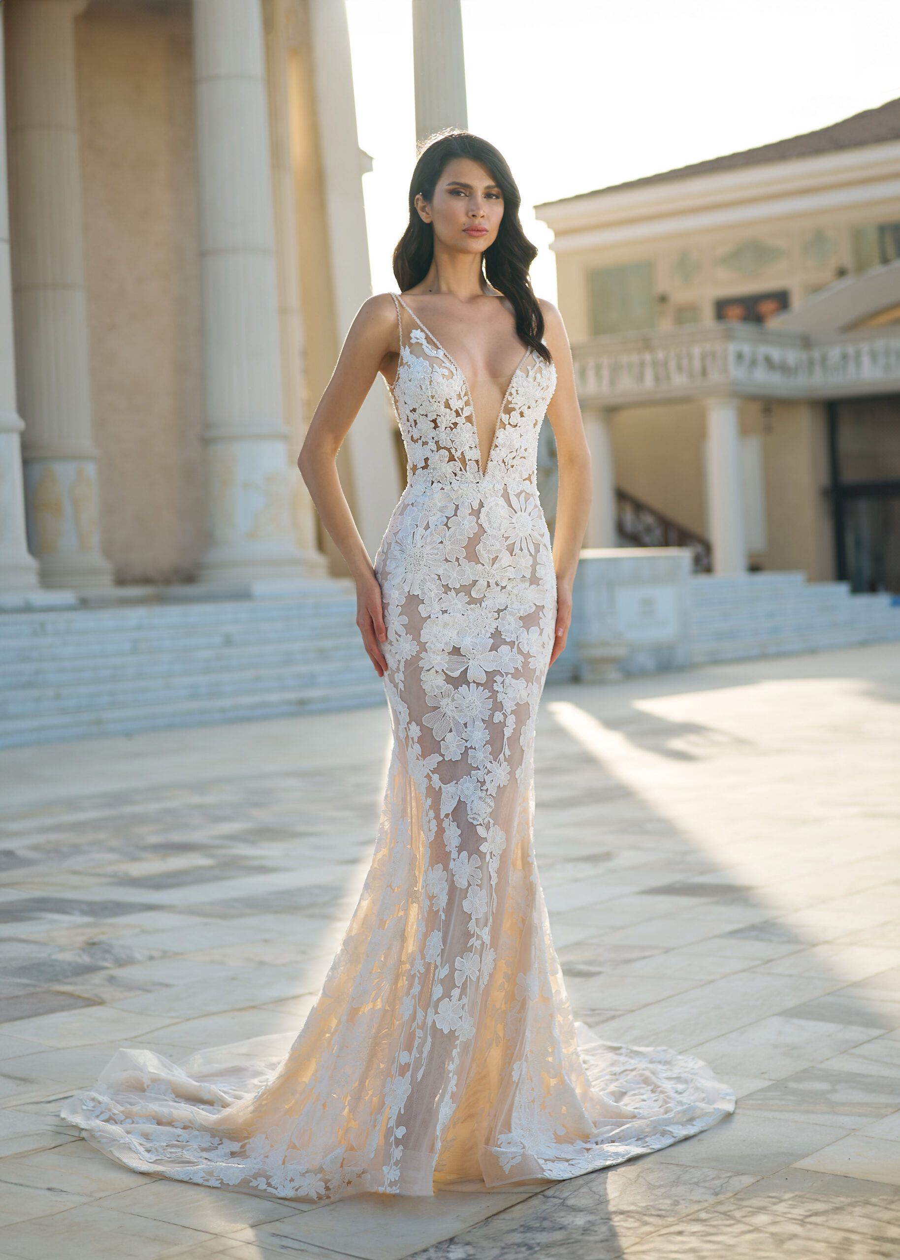 Woman in a white lace wedding dress standing on a beach at sunset.