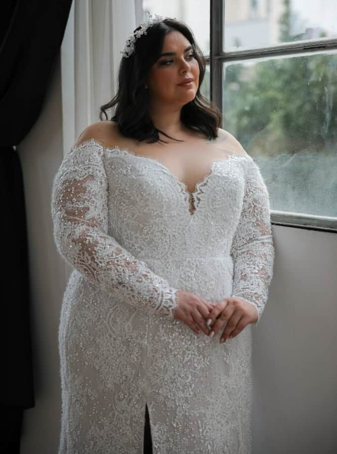 Woman in a white lace wedding dress standing on a beach at sunset.