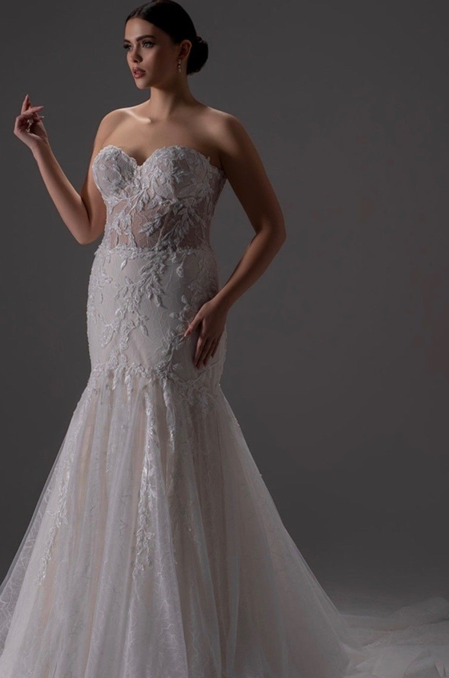 A blonde person wearing an off-the-shoulder, floor-length, white lace mermaid wedding dress against a plain gray background.