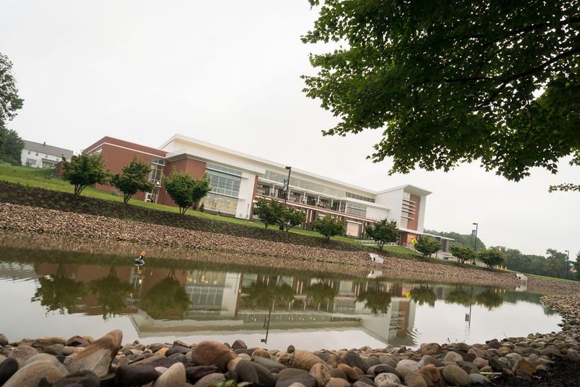 Modern campus building reflected in a pond, with rocks in the foreground and trees framing the scene.