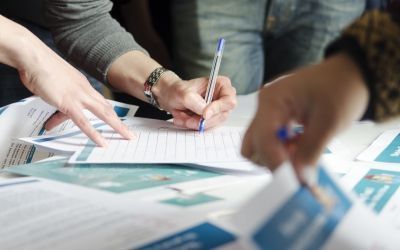 Hands writing on paperwork at a table with charts and documents spread out