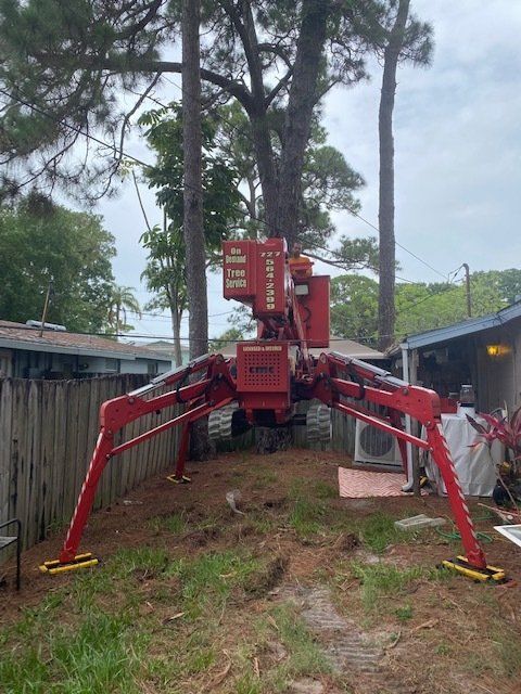 Red spider-like aerial lift on a grassy lawn; supports extended, near a wooden fence and trees.