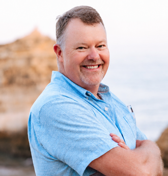 Smiling man in a light blue shirt standing with arms crossed near a rocky shoreline