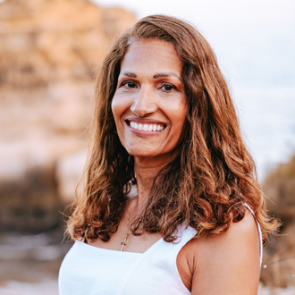 Smiling woman with long curly hair in a white tank top outdoors against a blurred rocky background