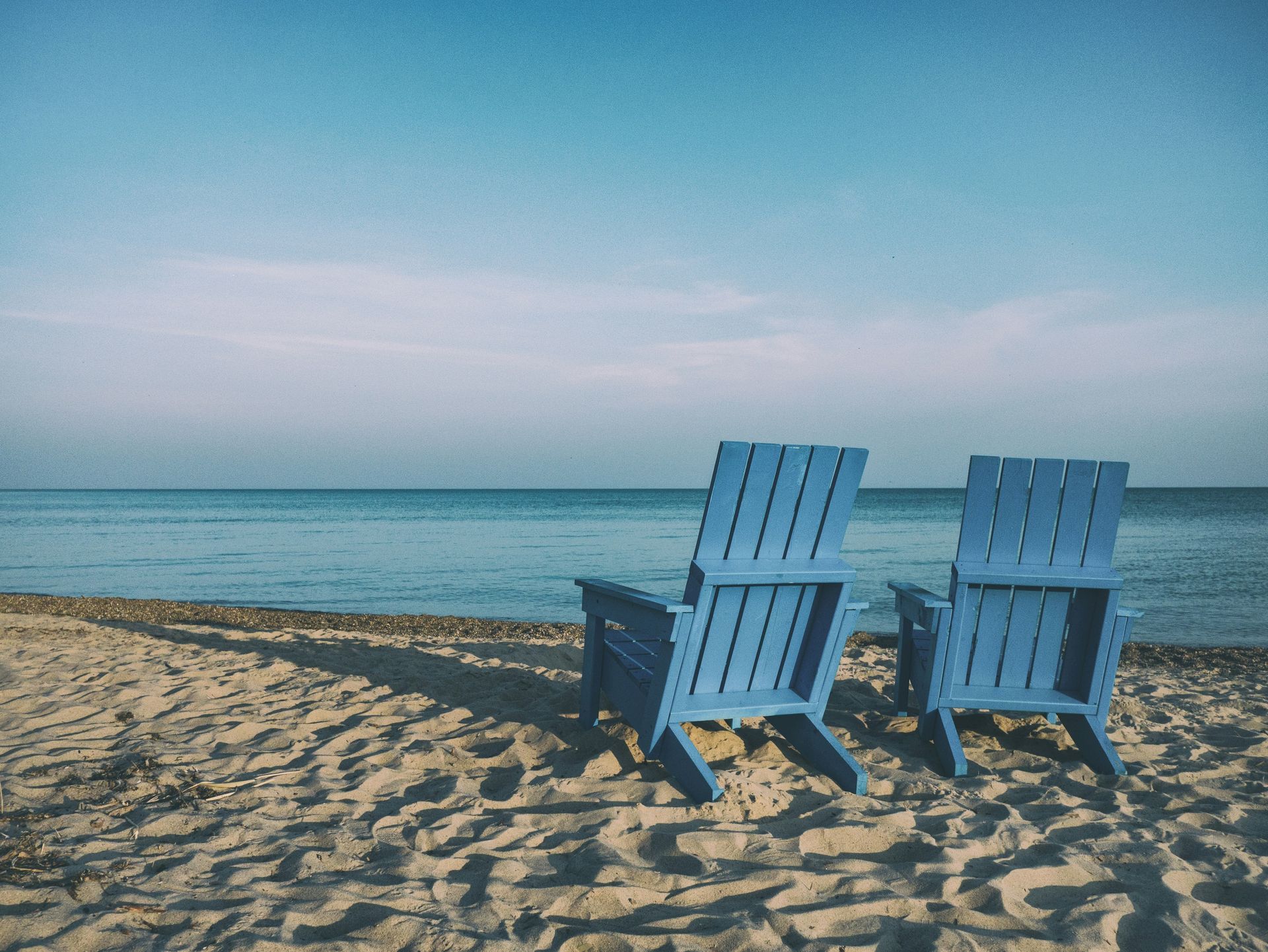 Two blue wooden chairs on a sandy beach facing a calm ocean at sunset