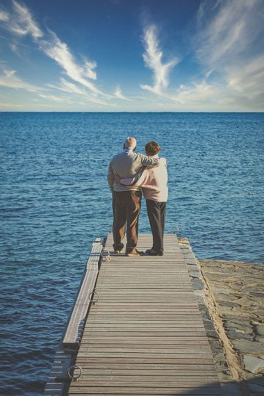 Two people stand on a wooden pier, looking out over a calm blue lake under a partly cloudy sky.