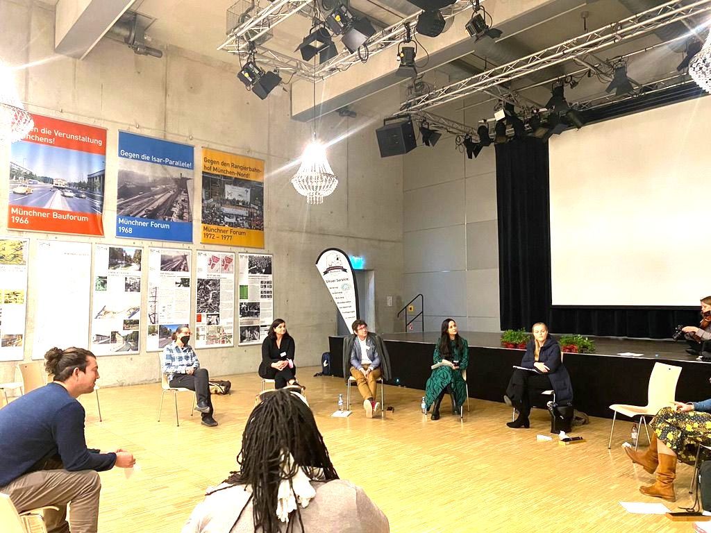 Sitzkreis in einem gorßen Saal, an der Wand Plakate und eine Projektionsleinwand