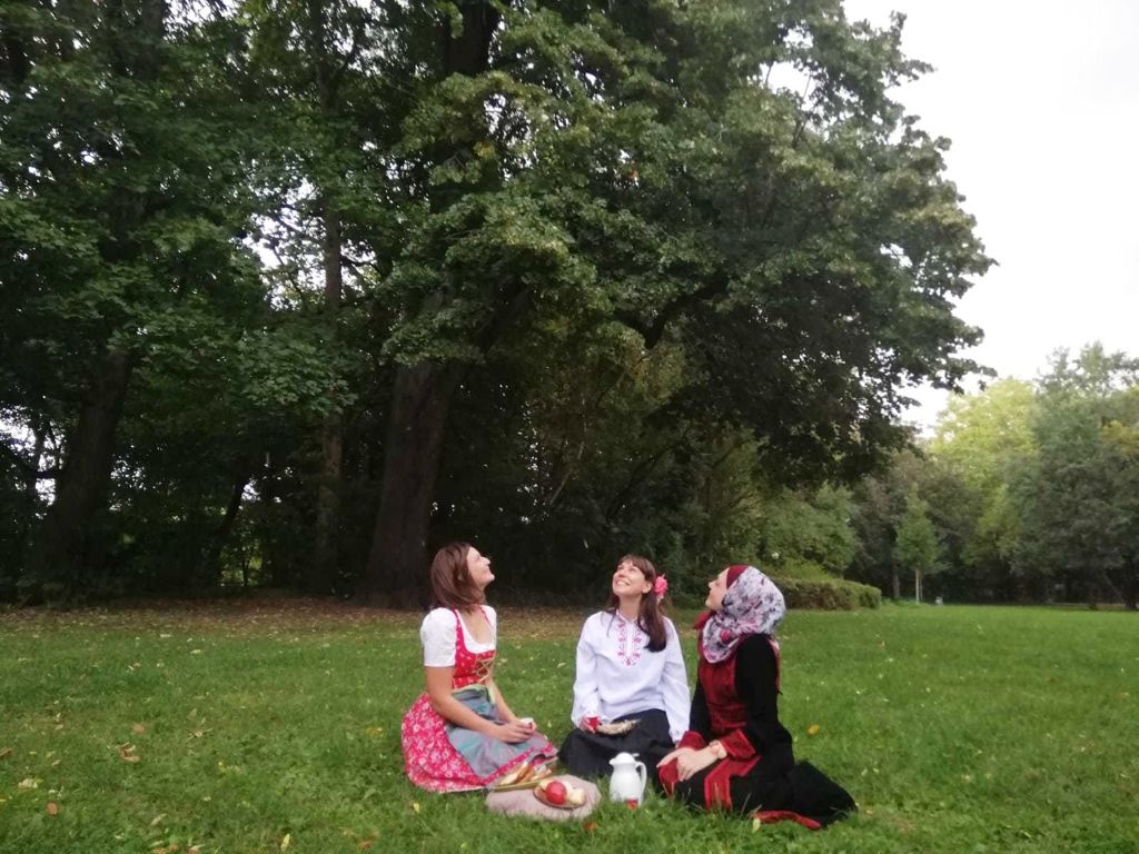 Drei jungen Frauen auf einer Wiese blicken in die Wolken, im Hintergrund Gebüsch