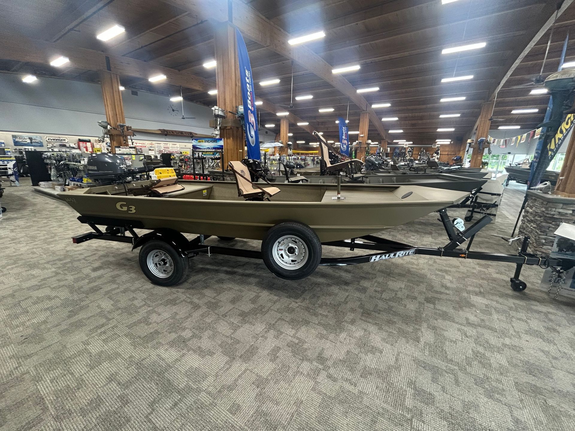 Small camouflage boat on a trailer inside a warehouse with rows of lights overhead