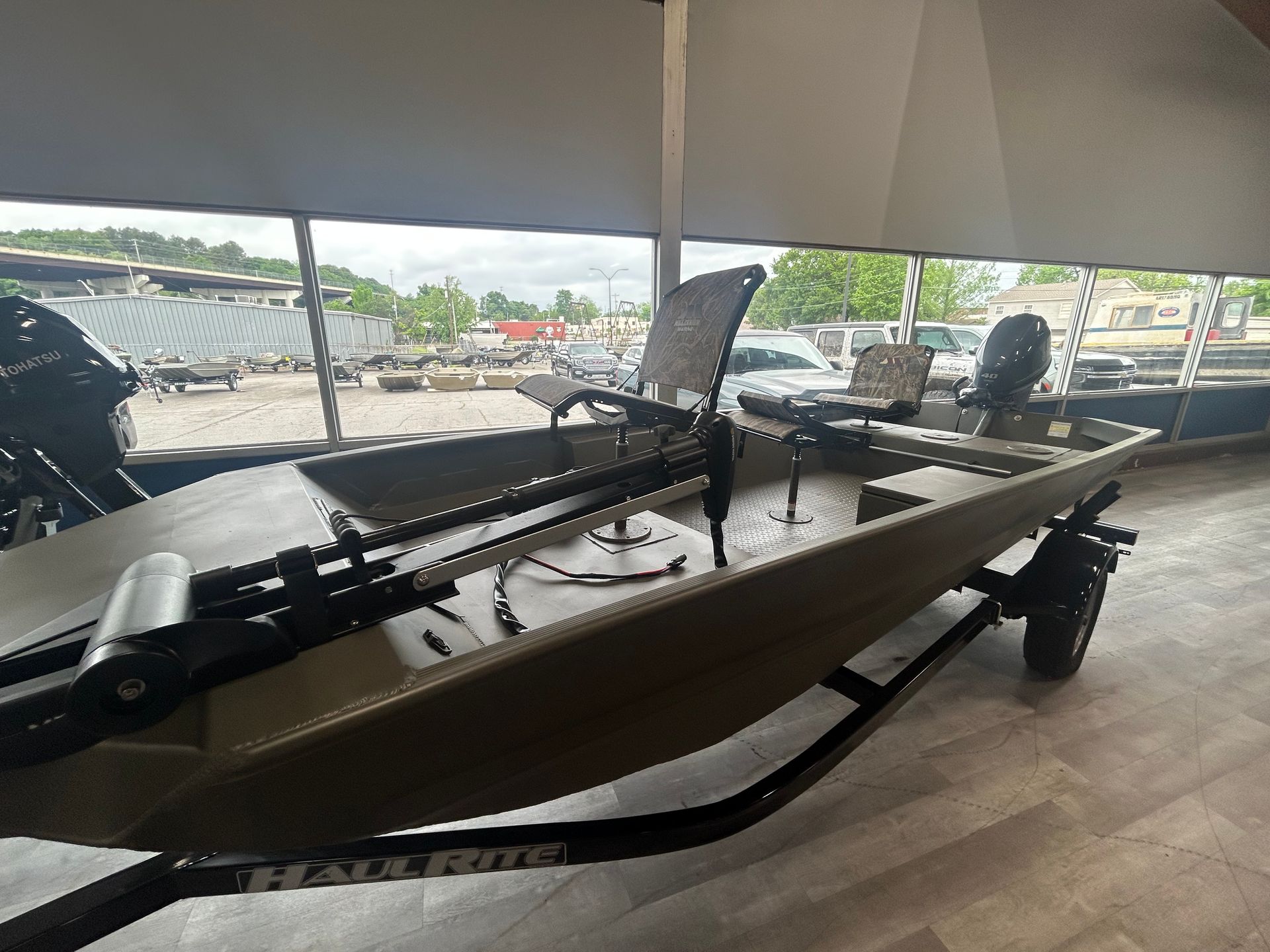 A tan aluminum fishing boat sits on a black trailer inside a showroom with large windows.