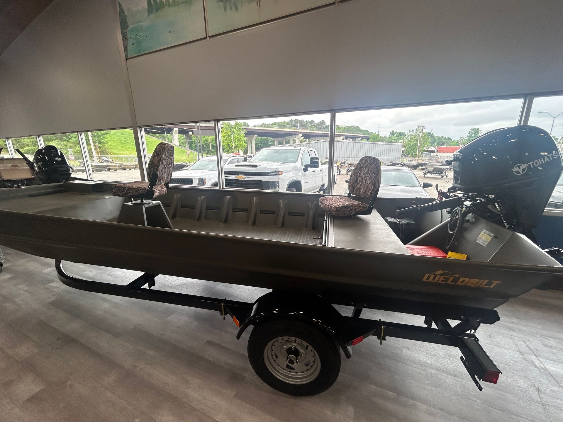 Black flat-bottom boat on a trailer under a covered area, with outboard motor and seating, near a parking lot