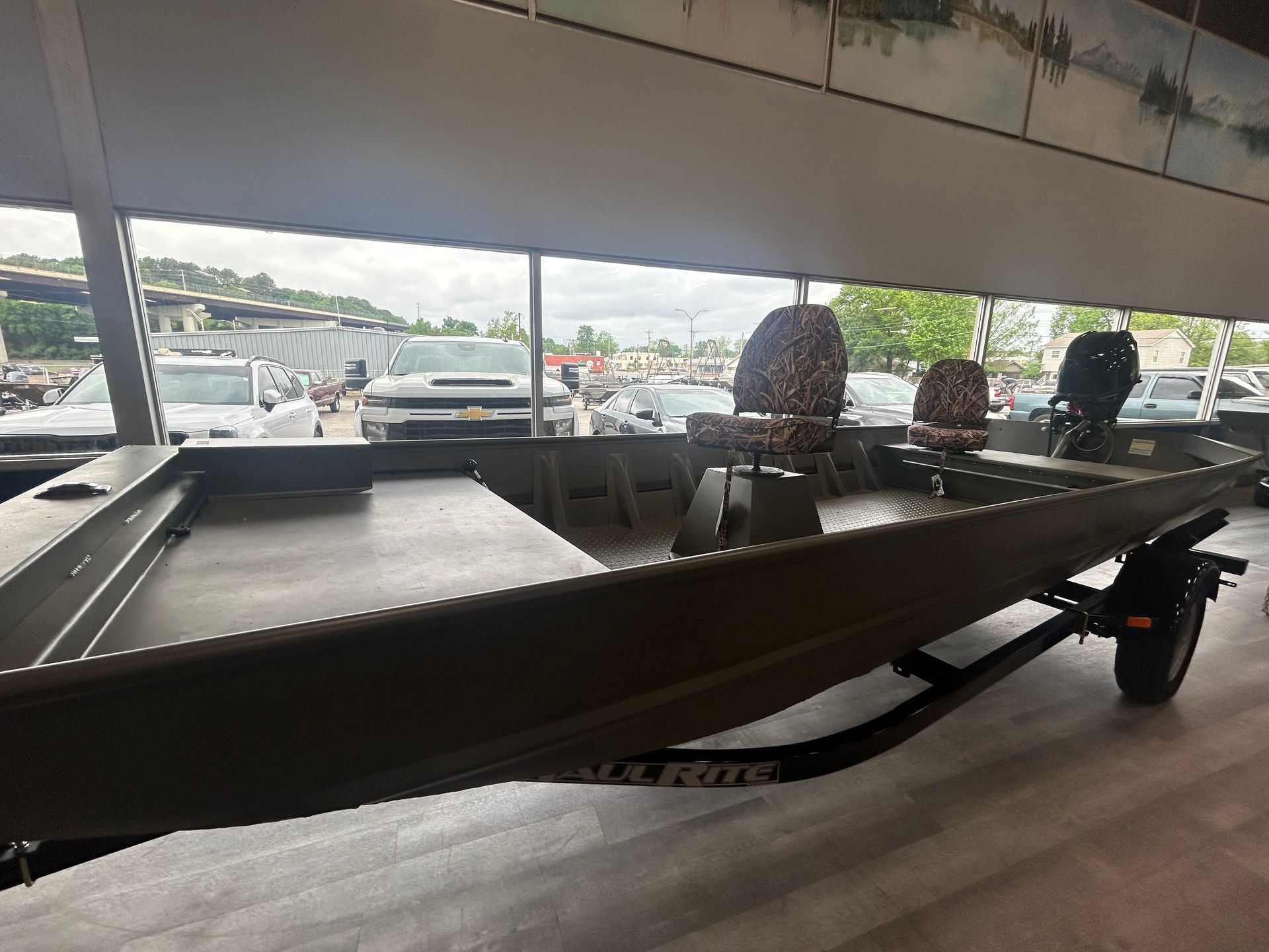 A dark green aluminum jon boat with camouflage seats and a front casting deck, sitting on a trailer in a showroom.