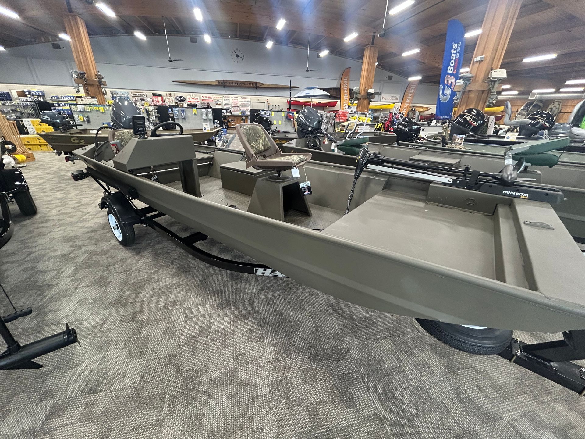 Gray flat-bottom fishing boat on stands in a large indoor showroom with other boats in the background