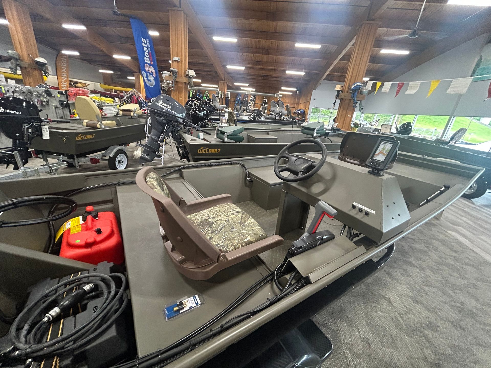 Inside a boat showroom, a small fishing boat with a steering wheel and seats is displayed indoors.