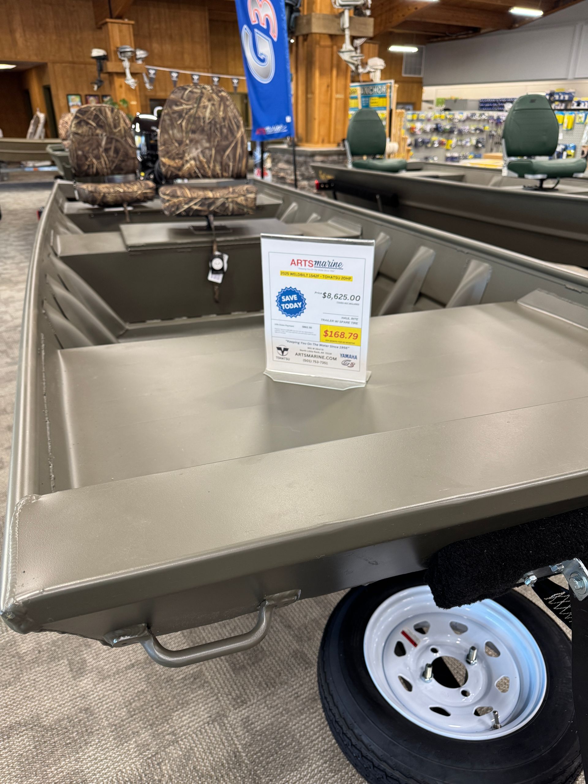 An olive-drab aluminum fishing boat with camouflage seats and a price tag sits in a showroom, parked on a trailer.