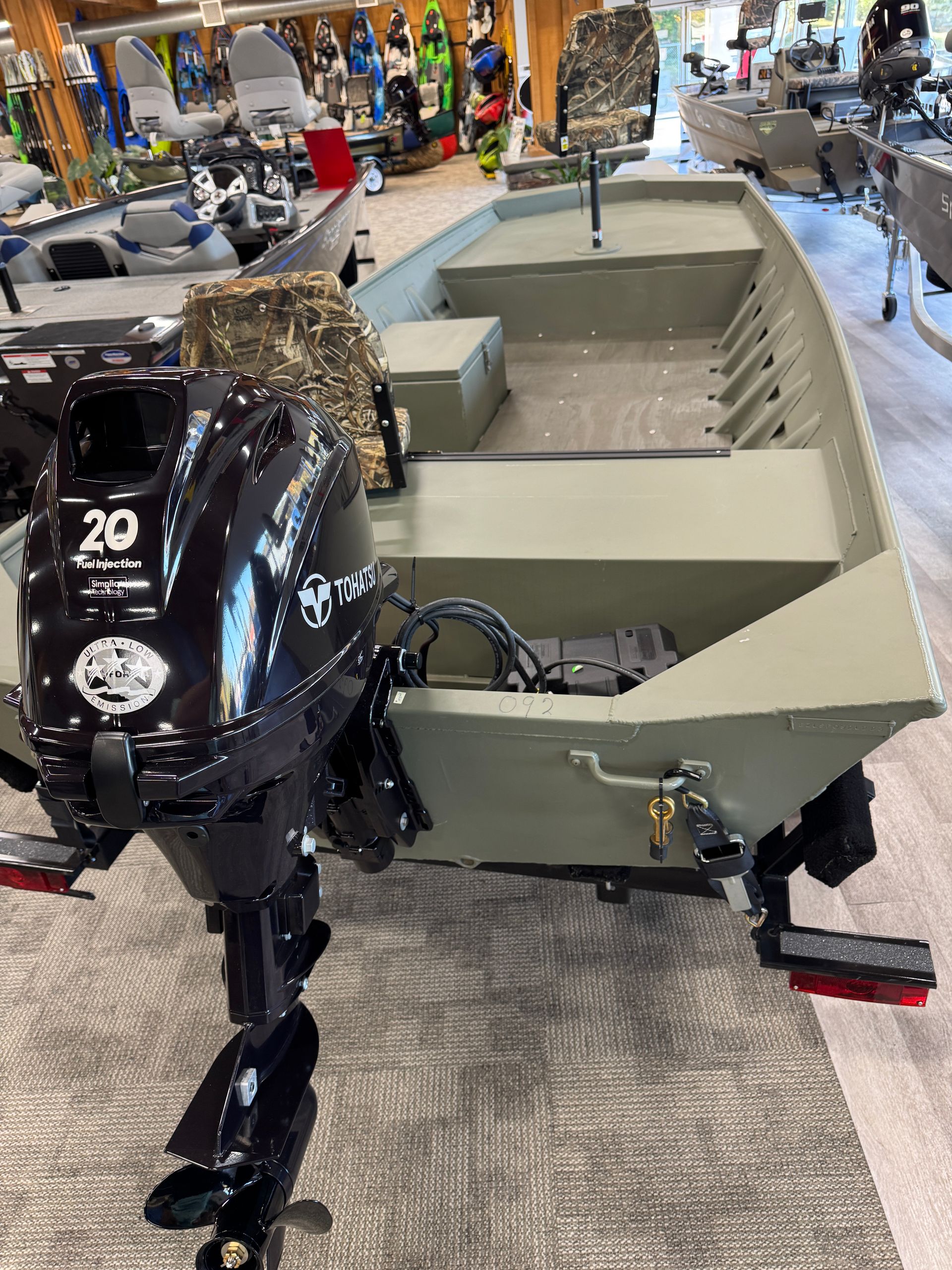 Olive-green pedal boat displayed indoors beside a black motor unit and camouflage seat cover