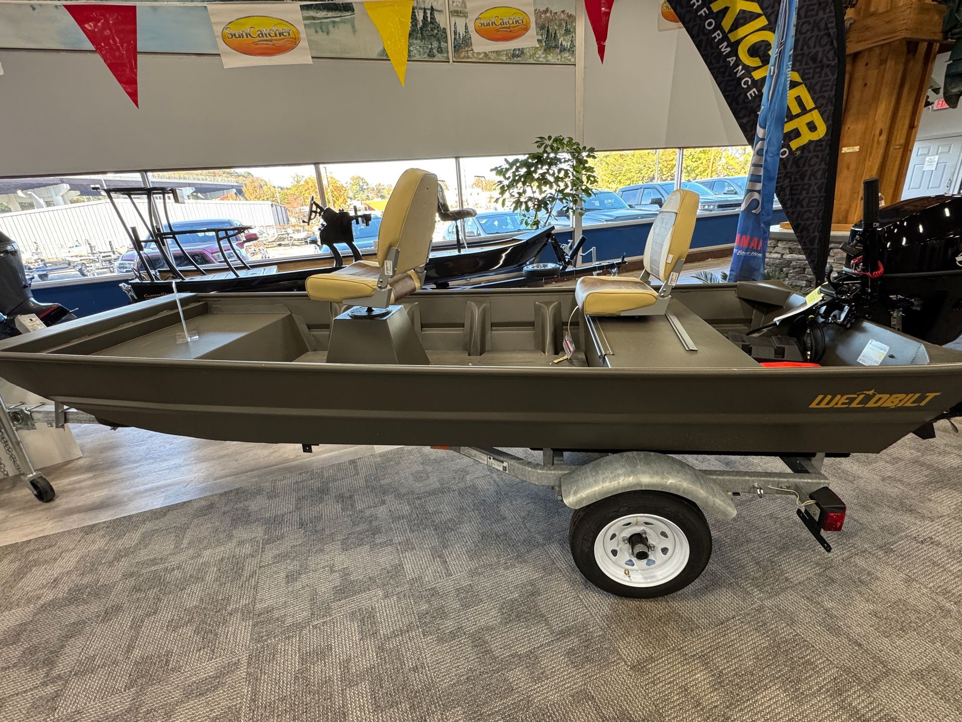 A small, olive-colored aluminum boat on a trailer, with two tan seats, sits indoors.