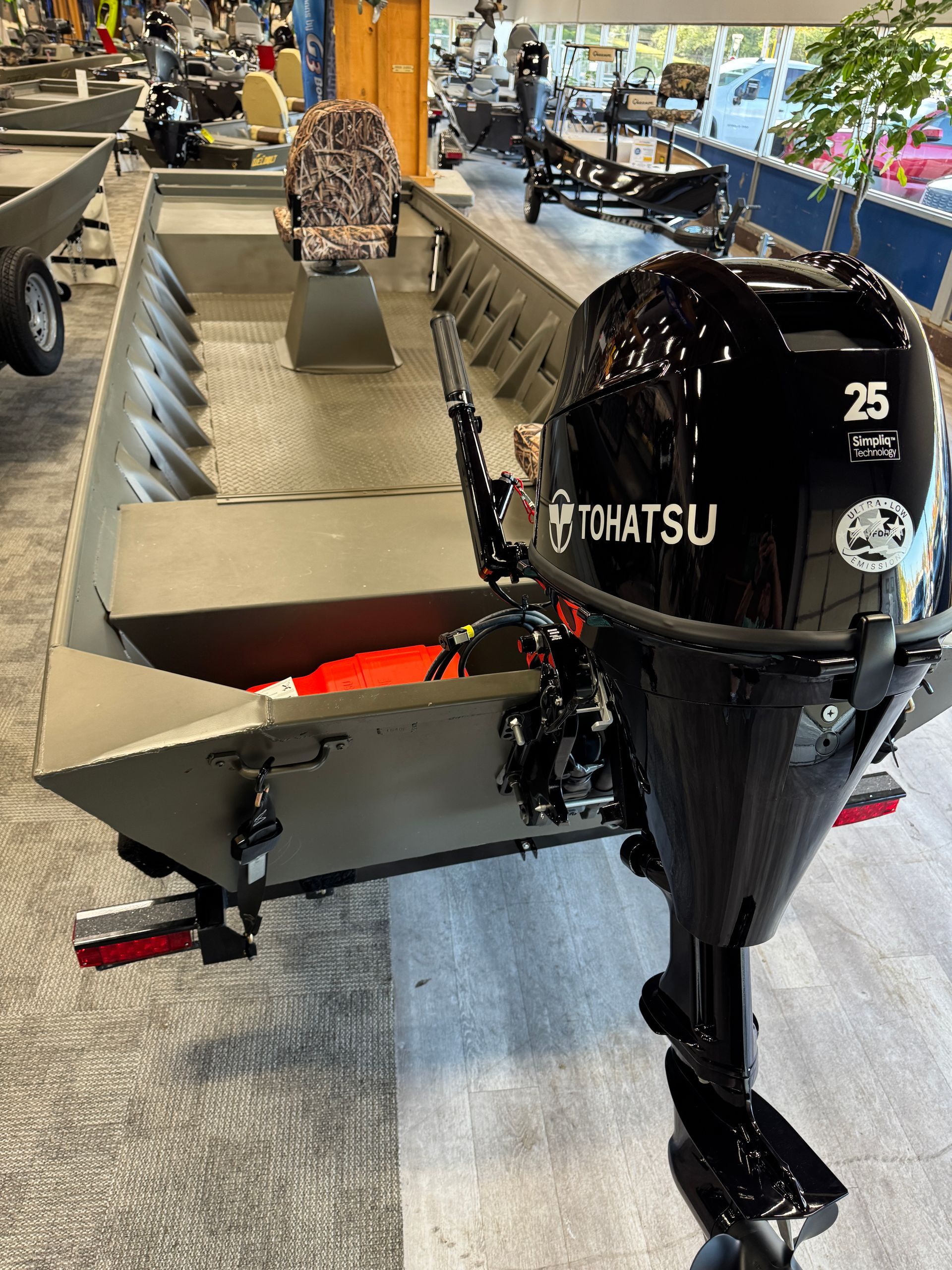 Small aluminum boat with a black Tohatsu outboard motor, shown indoors on a showroom floor.