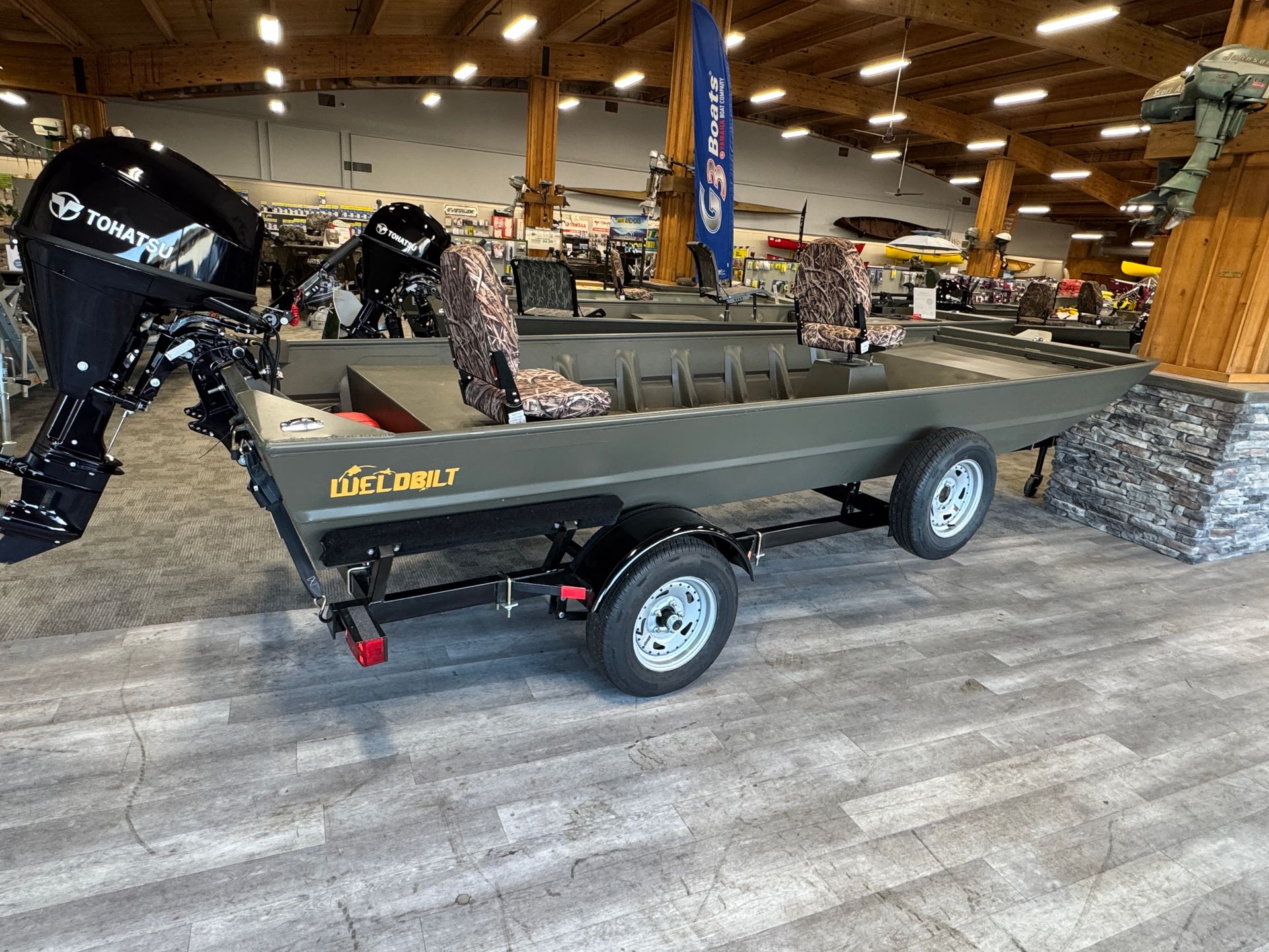 Green jon boat on a trailer, with outboard motor and camo seats, inside a shop.