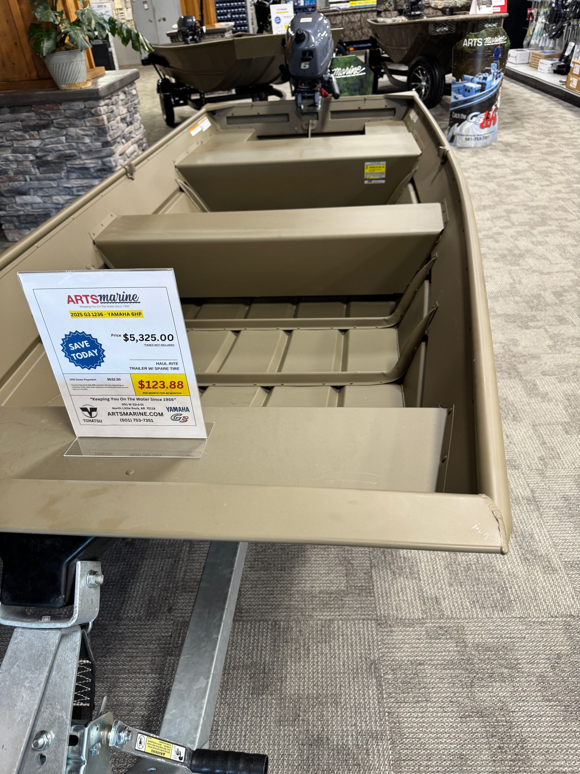 Tan boat with bench seats and a sale sign displayed indoors on carpeted showroom floor
