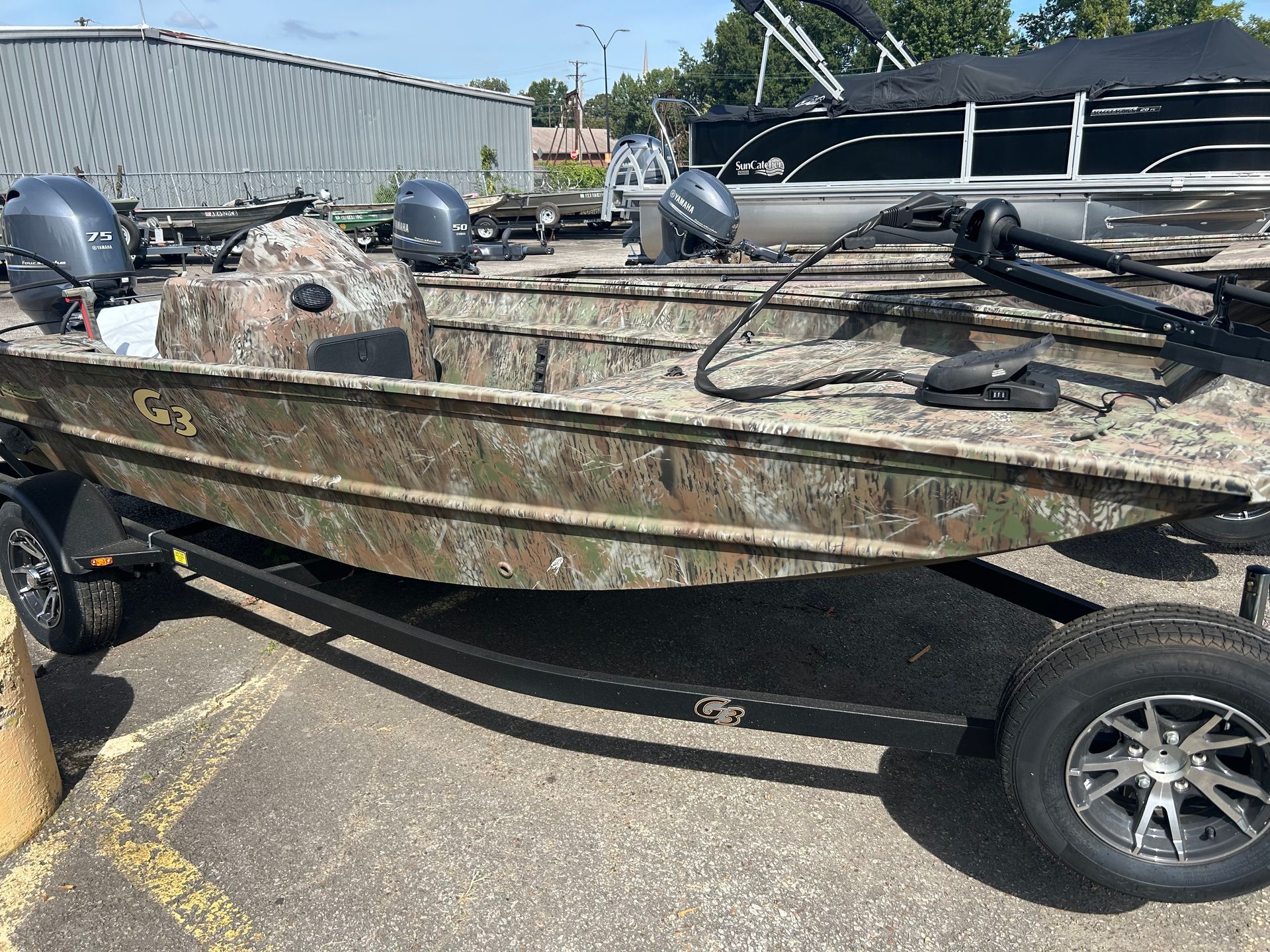 Camouflaged flat-bottom boat on a trailer in a parking lot, with fishing gear on deck
