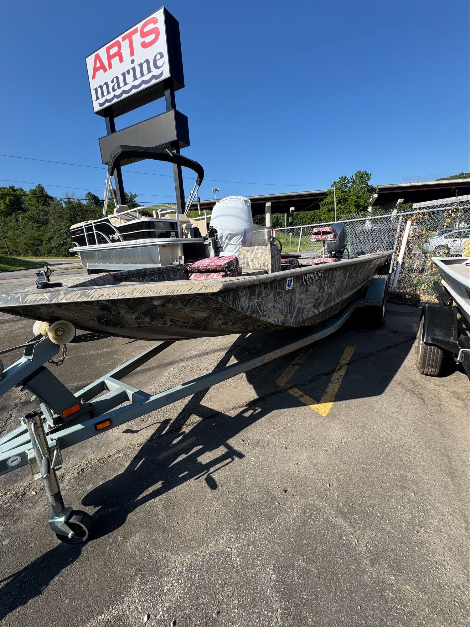 Camouflage-painted boat on trailer in parking lot, under a sign for Arts Marine.