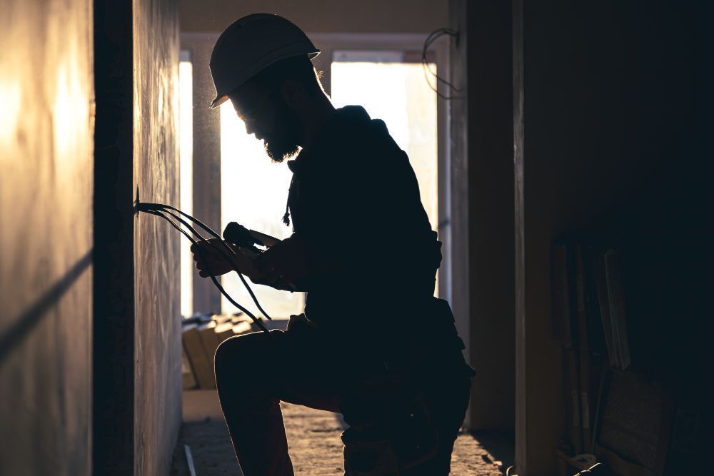 Silhouetted worker kneeling in a dim doorway, wiring a wall with tools in hand.