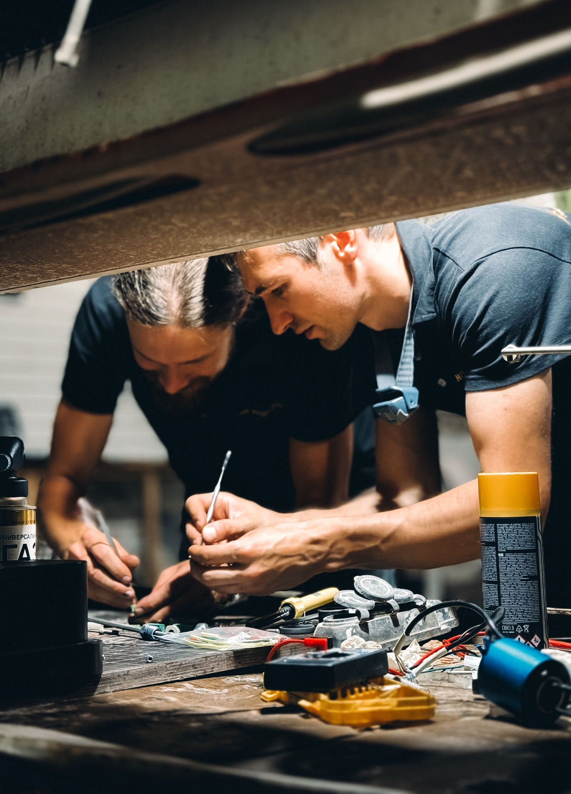 Two technicians crouch under a structure, wiring equipment on the floor with tools and cables.