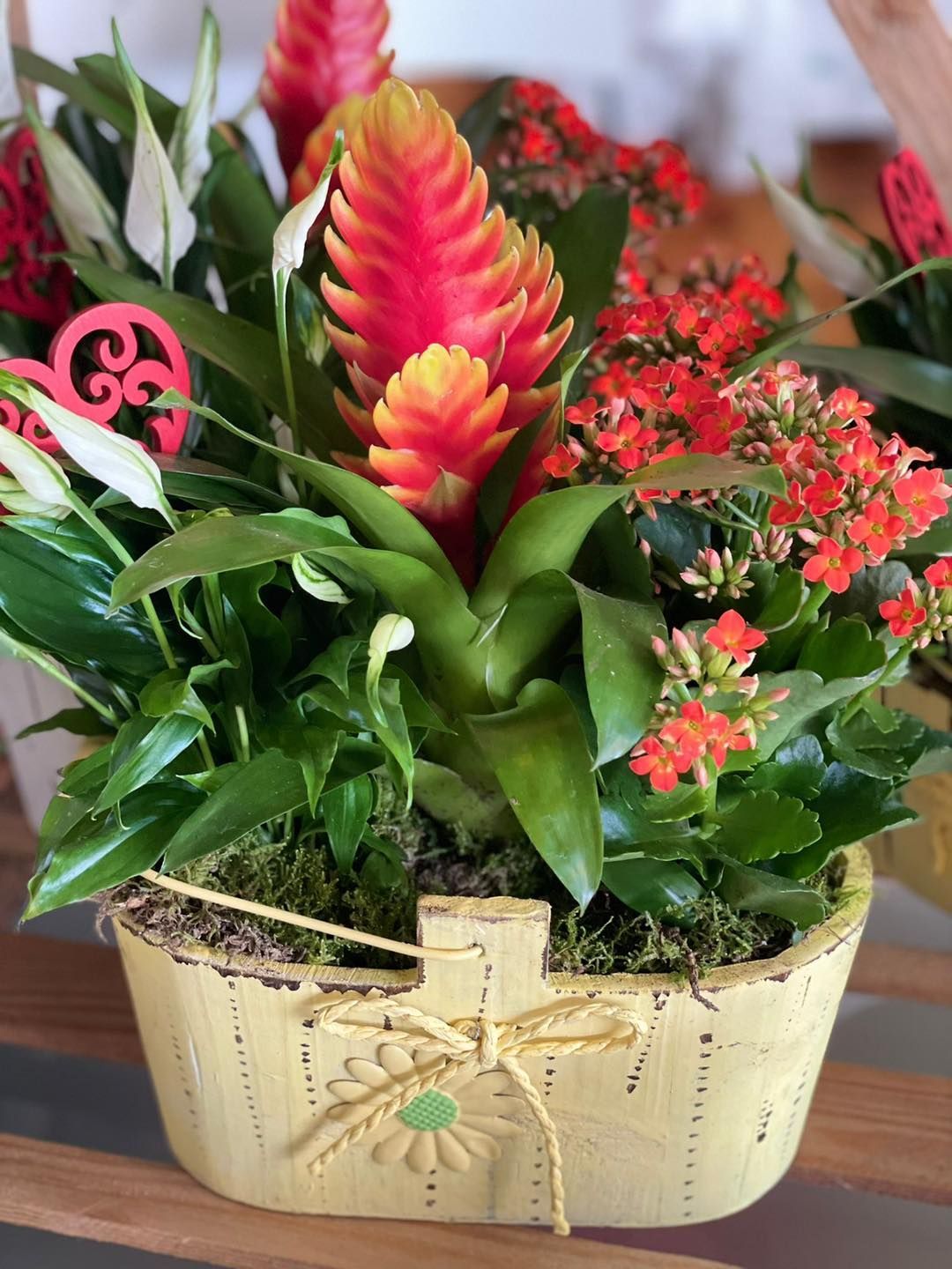 Composizione floreale con fiori rossi e arancioni e fogliame verde in un cestino intrecciato color crema con fiocco di nastro.