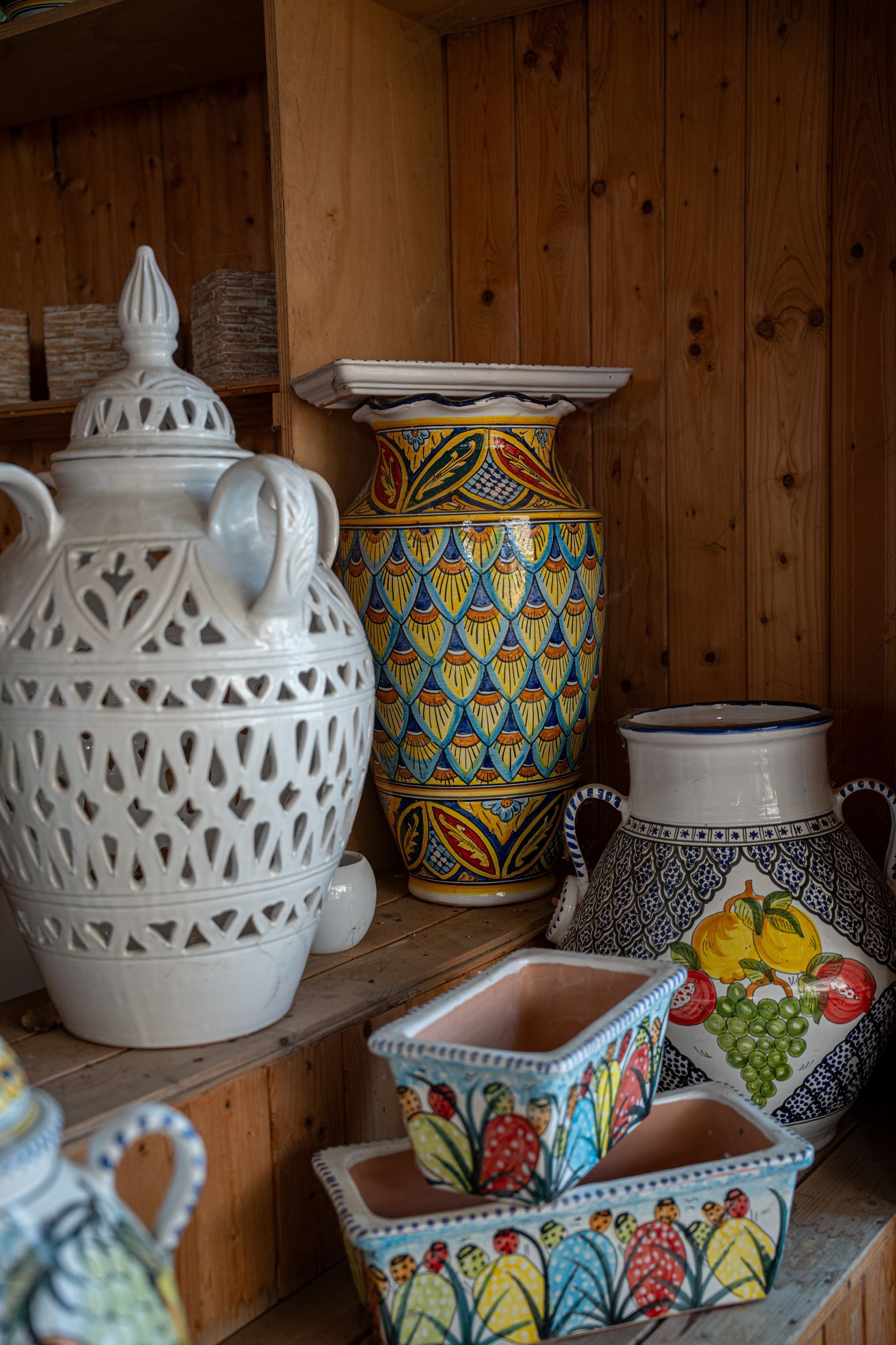 Assortimento di vasi e giare in ceramica colorata su una mensola di legno, tra cui pezzi in stile marocchino con decorazioni in pizzo bianco e altri motivi decorativi.