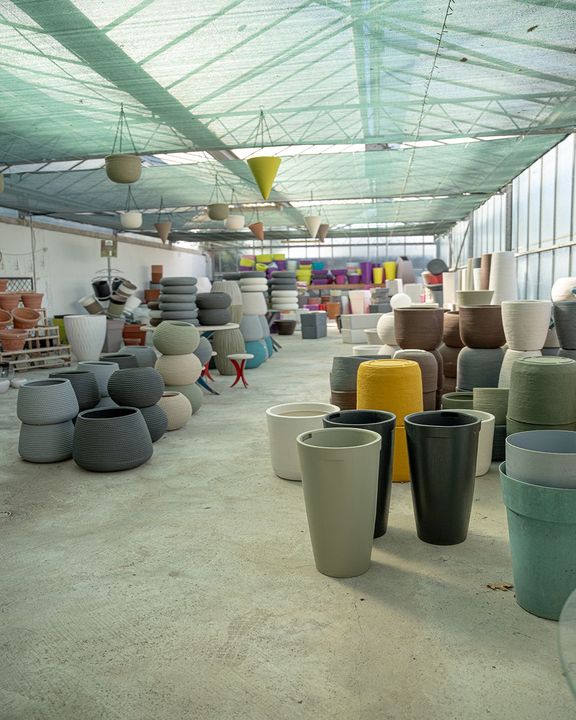 Showroom all'aperto con sgabelli intrecciati colorati e fioriere sotto una tettoia verde ombreggiante.