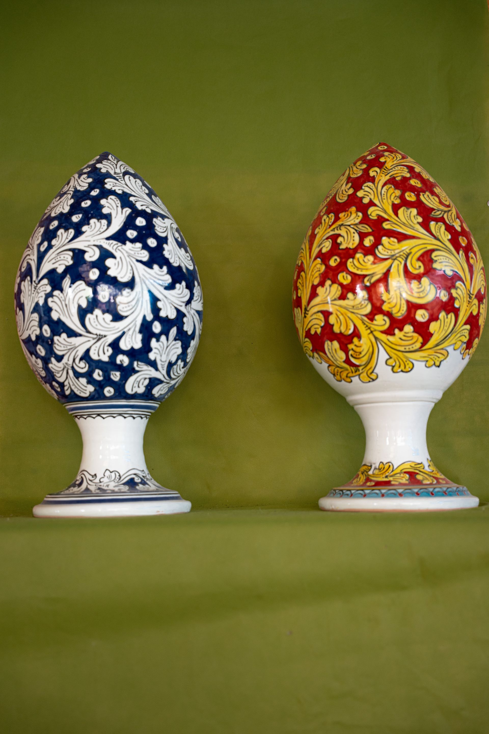Due elaborati oggetti decorativi a forma di uovo, uno blu e bianco, l'altro rosso e oro, su sfondo verde.
