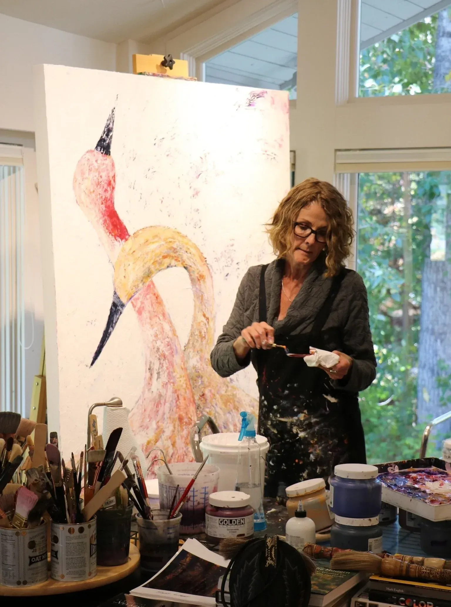 Woman paints in a studio. Large canvas features abstract flamingo art. Paint on apron and table, natural light.