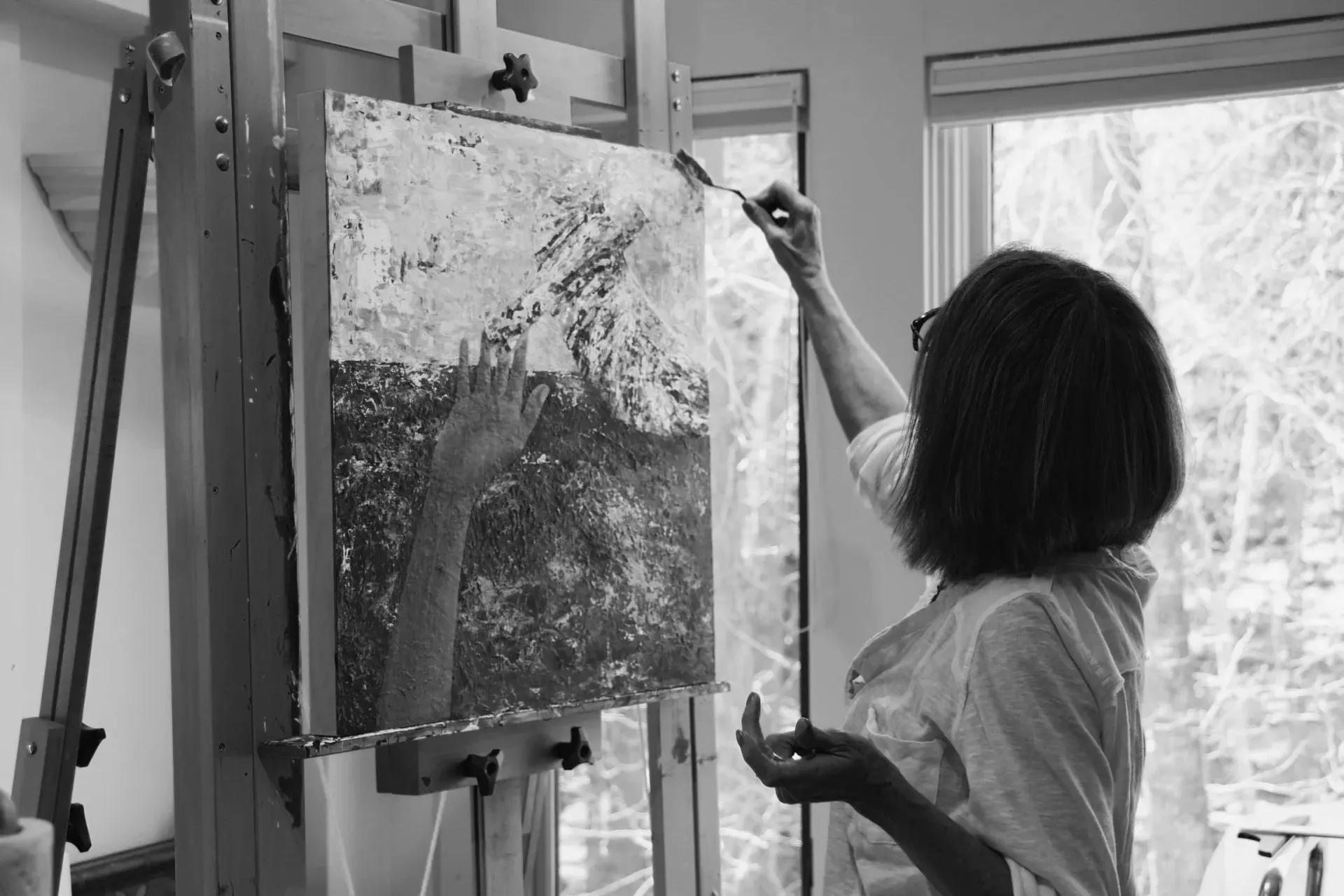 Woman painting at easel, reaching towards canvas with brush. Window in background.