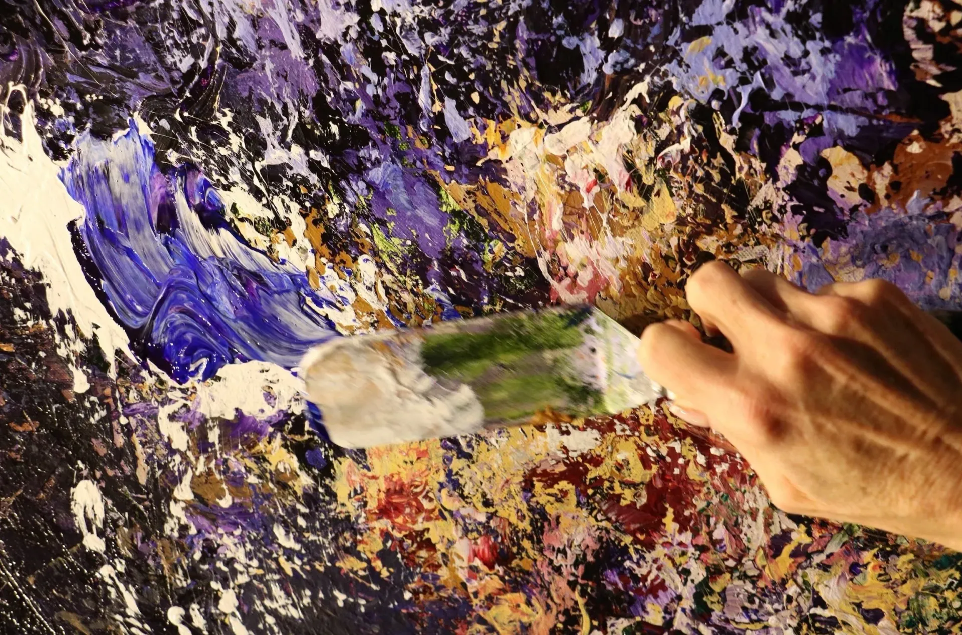 Hand using a palette knife to spread blue and green paint on a canvas with other colors.