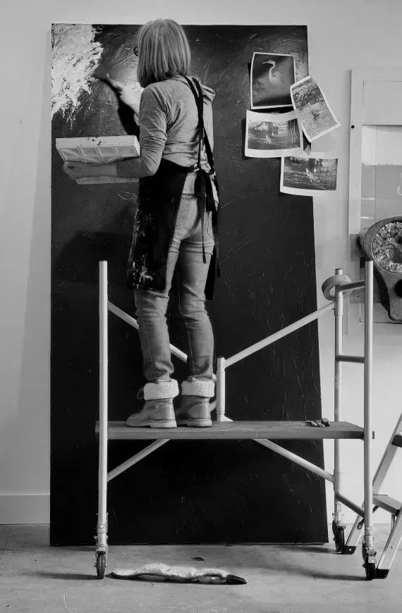 Artist standing on a small platform painting a large canvas in a studio. Black and white.