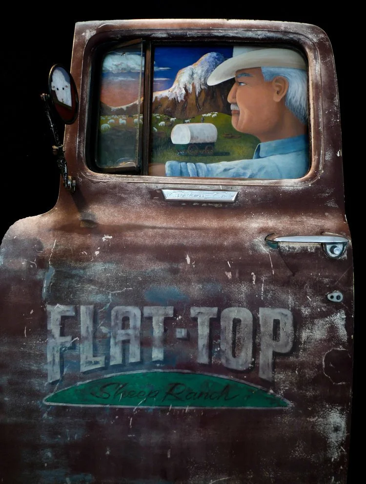 Rustic truck door with driver wearing a cowboy hat. Mountains visible through the window. 