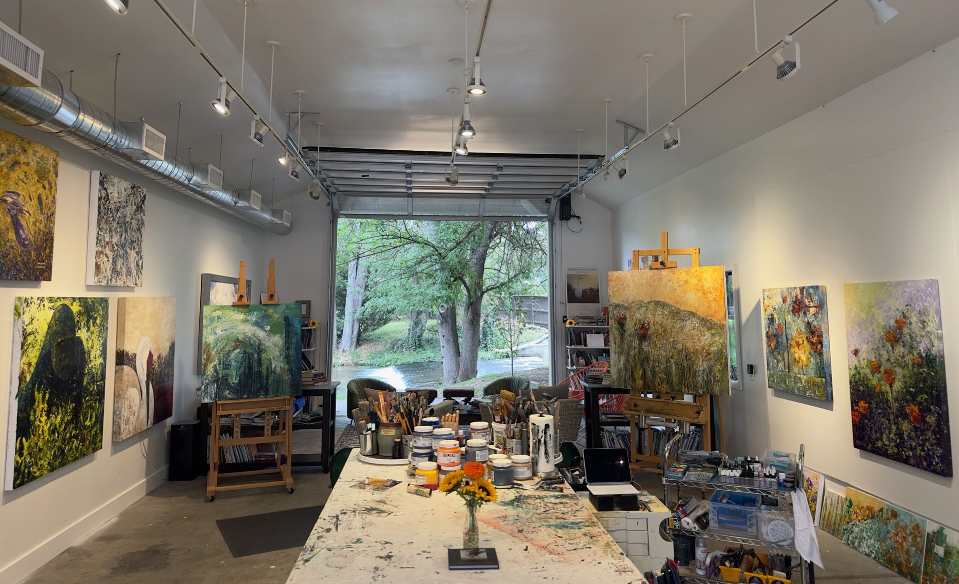 Art studio with paintings on the walls and easels.  Open door reveals trees and a table with paint supplies.