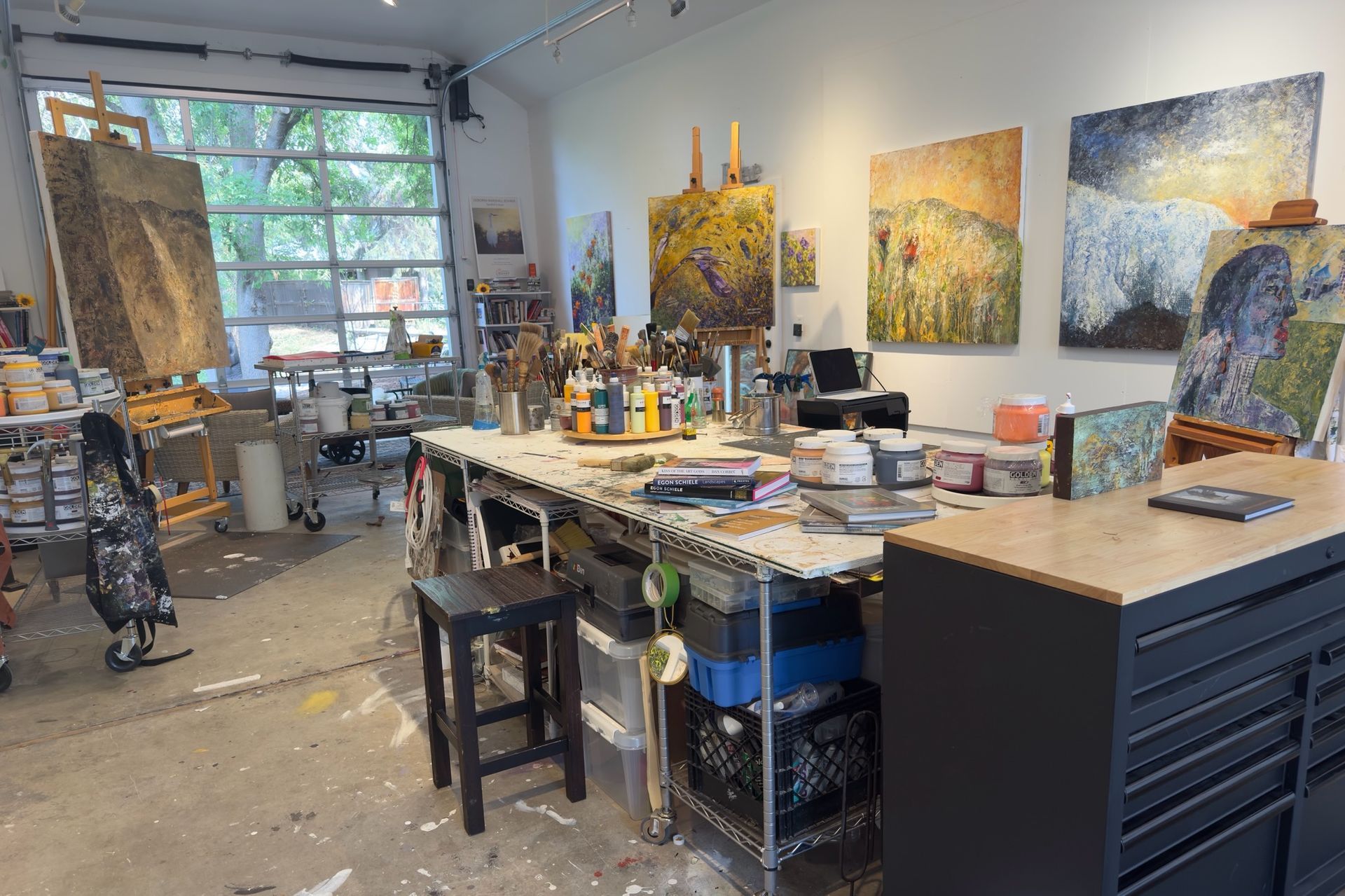 Artist's studio with paint-splattered floor, art supplies, worktable, and paintings on the wall.