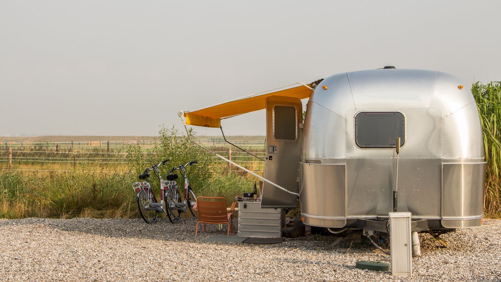 Silver camper with awning, bicycles, and chair set up on a gravel lot next to a grassy field.