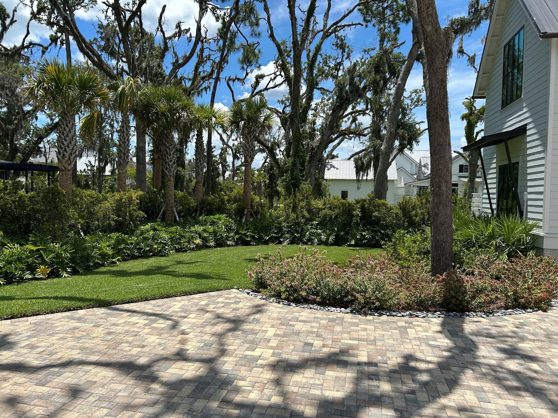 Brick patio with green lawn and landscaping, trees, and a white house under a bright blue sky.
