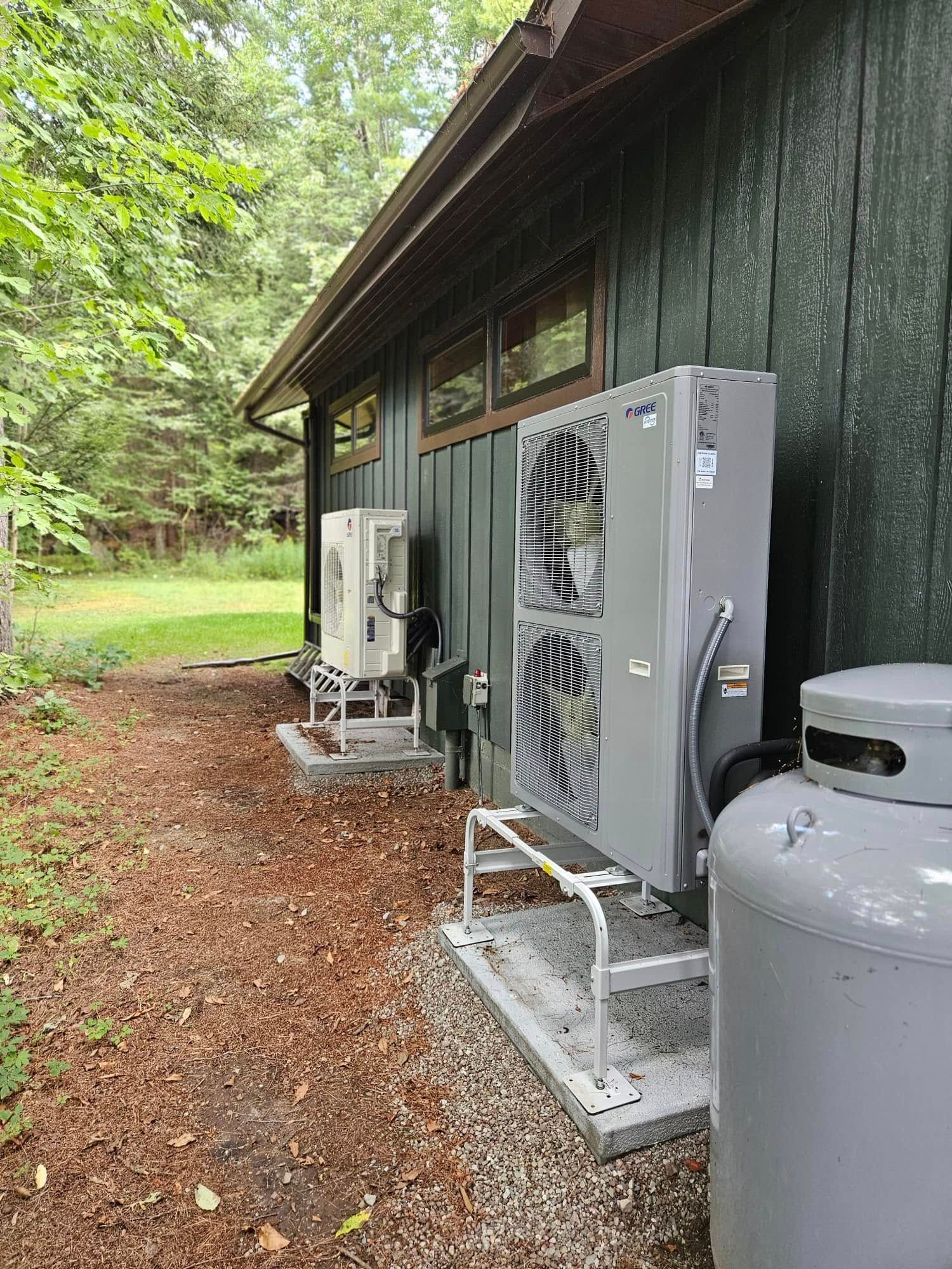 Outdoor HVAC units beside a dark wooden building, with a gray propane tank in the foreground.