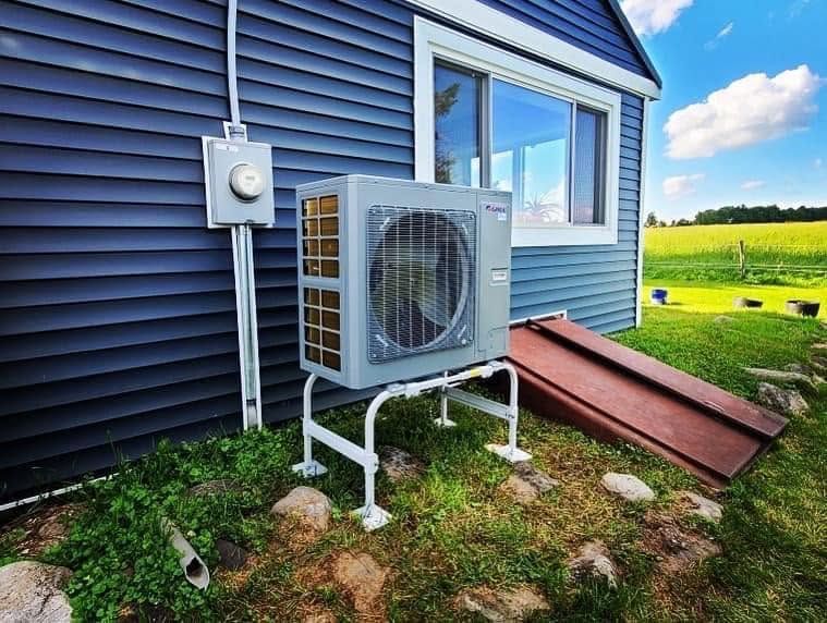 Outdoor air conditioner unit beside a blue house, near a window and ramp in a grassy yard